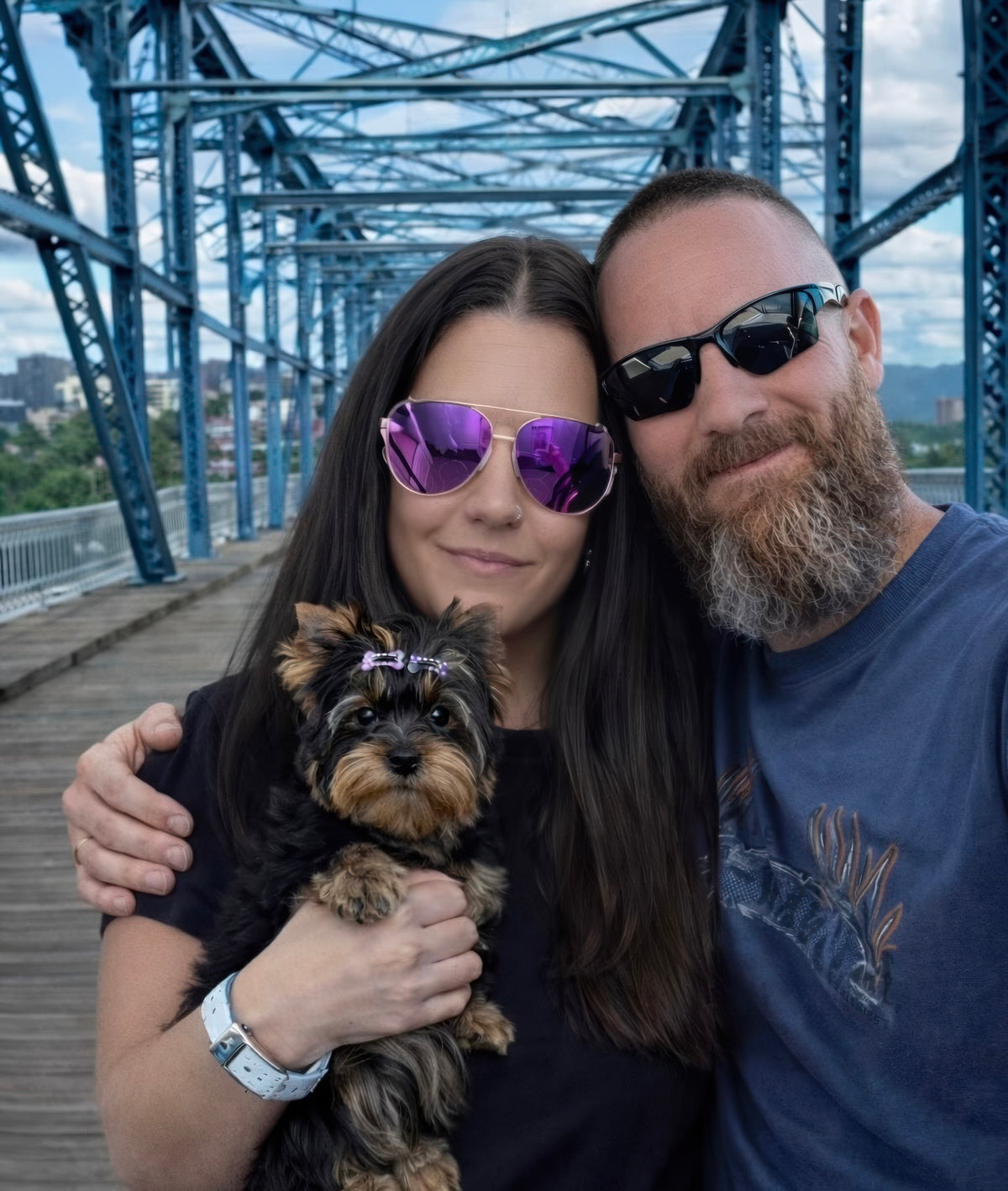 A couple wearing sunglasses and a woman holding a small Yorkshire Terrier dog on a wooden boardwalk bridge with a blue steel truss structure in the background.