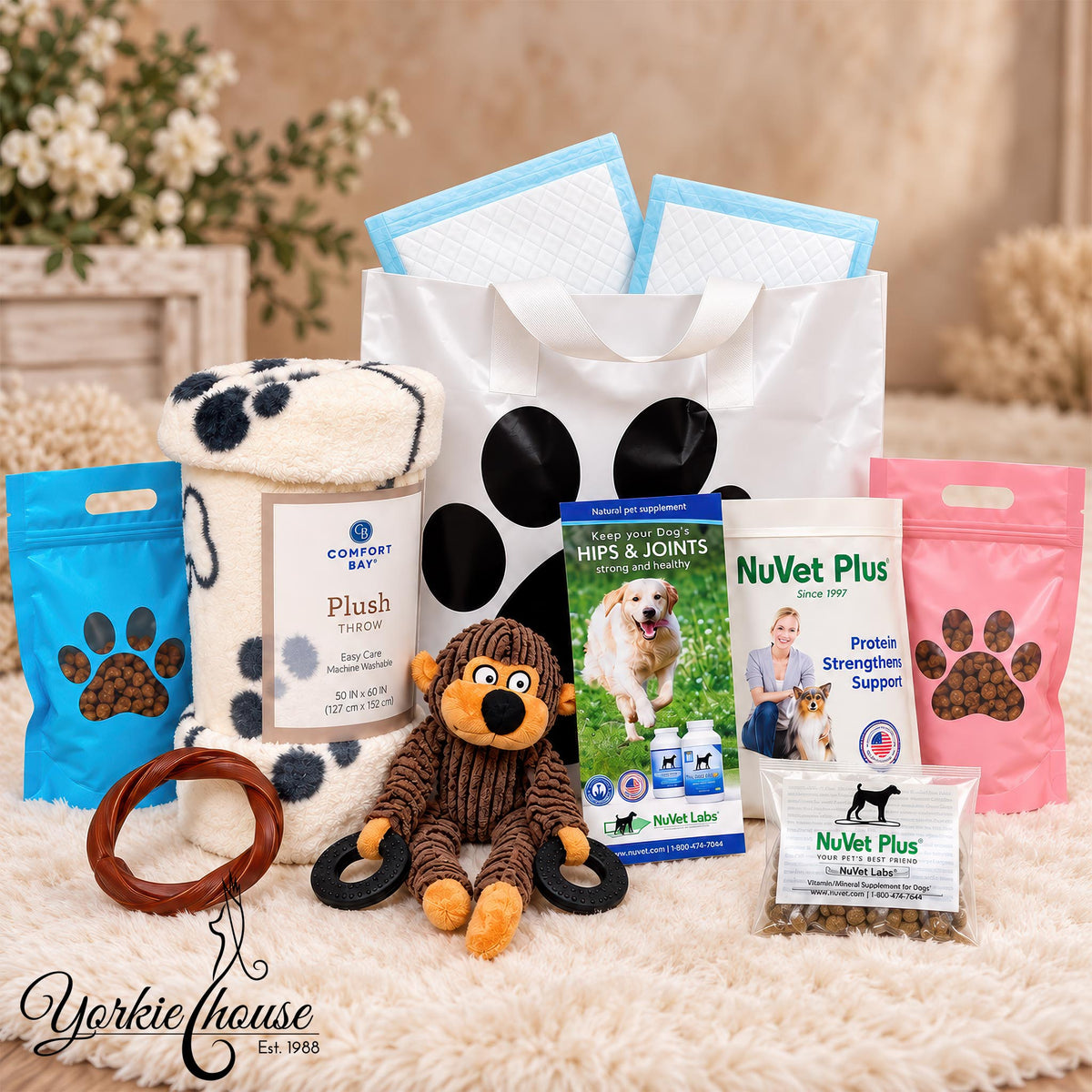 A curated pet care gift set arranged on a white faux-fur surface with a soft beige background. The set includes a large white tote bag with black paw prints, blue and pink dog treats, a plush toy, a chew treat bag, a lick mat, and several branded supplement packets including Comfort Kit, NuVet Plus, and Pet Care Labs.