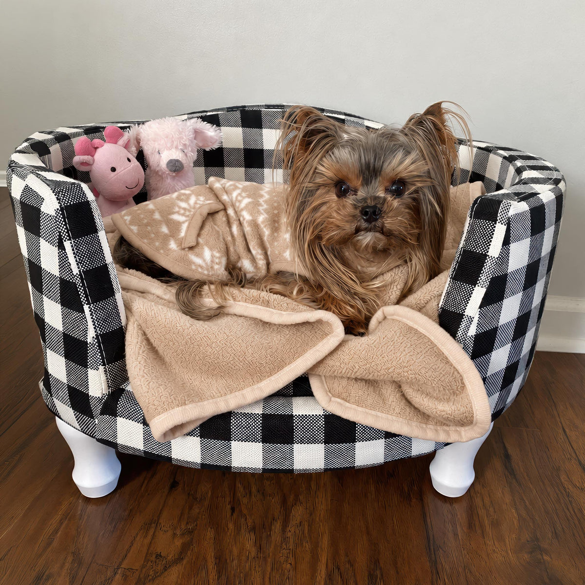 A small Yorkshire terrier dog sits in a round pet bed with a black and white buffalo plaid fabric, a beige fleece blanket, and two pink plush toys, set on a dark wood floor against a light wall.