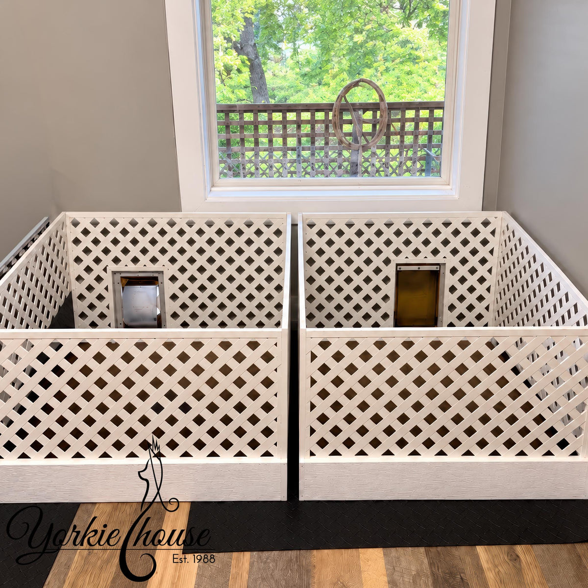 Two white lattice pet playpens with secure door compartments, positioned side-by-side in front of a window showing a green outdoor view. A black floor mat is placed underneath, and the Yorkie House logo is visible in the bottom left corner.