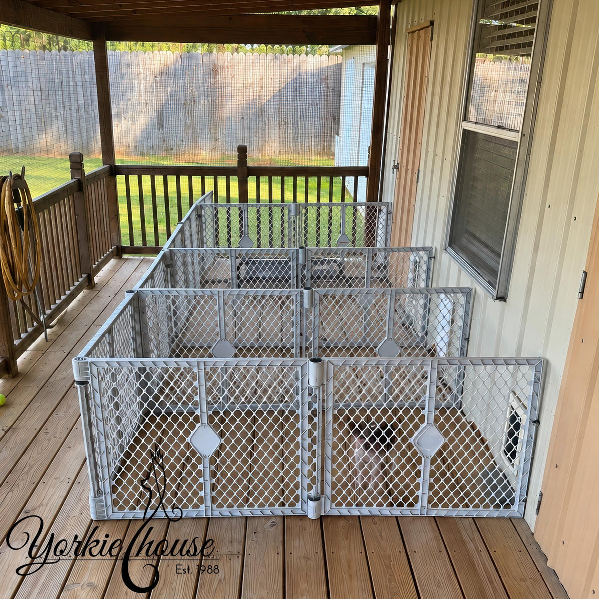 A set of silver metal chain-link pet playpens arranged on a wooden deck porch next to a house, with a wooden fence and yard visible in the background. A 'Yorkie House' logo watermark is in the bottom left corner.