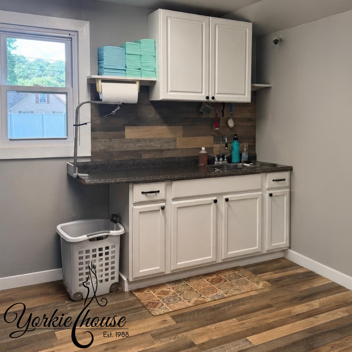 A laundry room corner with white cabinets, a dark countertop, a wood-look backsplash, a window, a hanging faucet, stacked mint towels, and a laundry basket on a wood floor. The Yorkie House logo appears in the lower left.